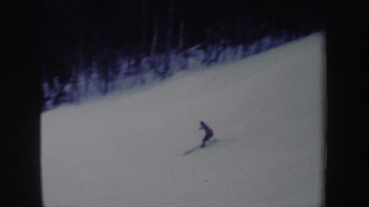 1962 New York home-movie frame showing the red-jacketed skier leaning into a downhill turn on a snow-covered slope.