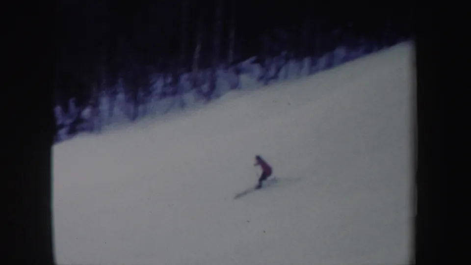 1962 New York home-movie frame showing the red-jacketed skier leaning into a downhill turn on a snow-covered slope.