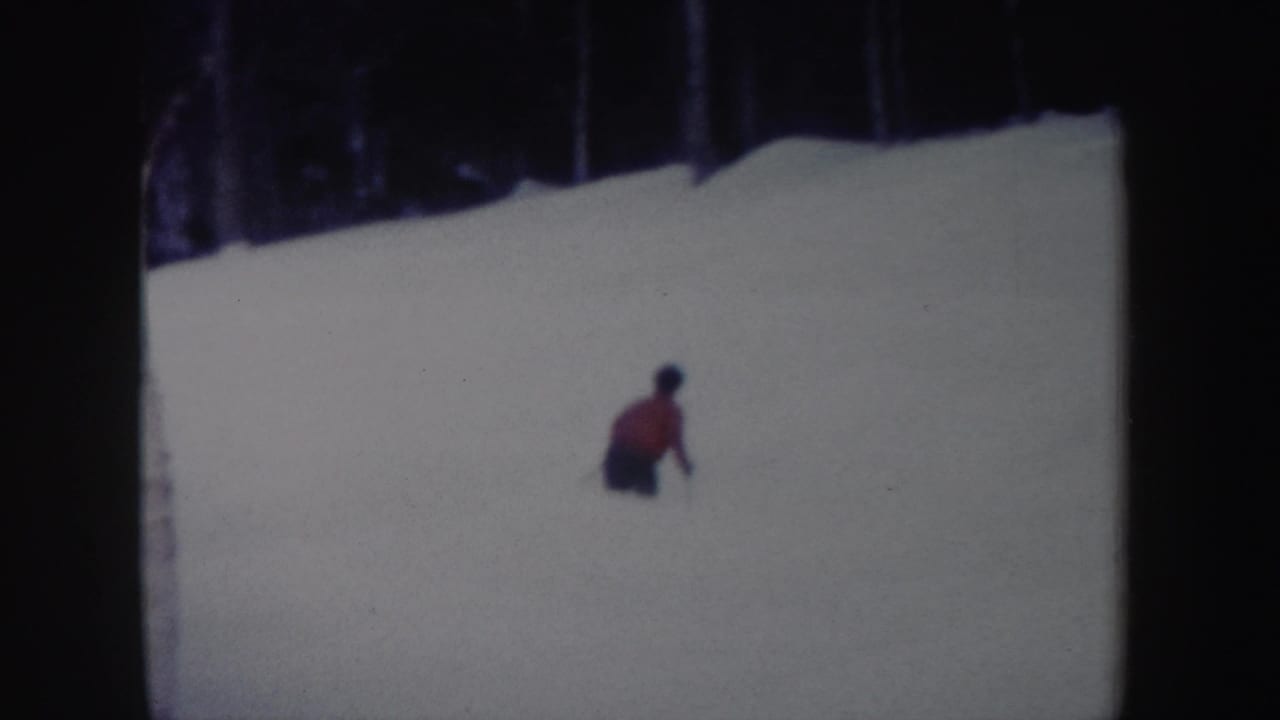 A red-jacketed skier crouches into a turn on the snowy New York slope in soft 8mm color footage.