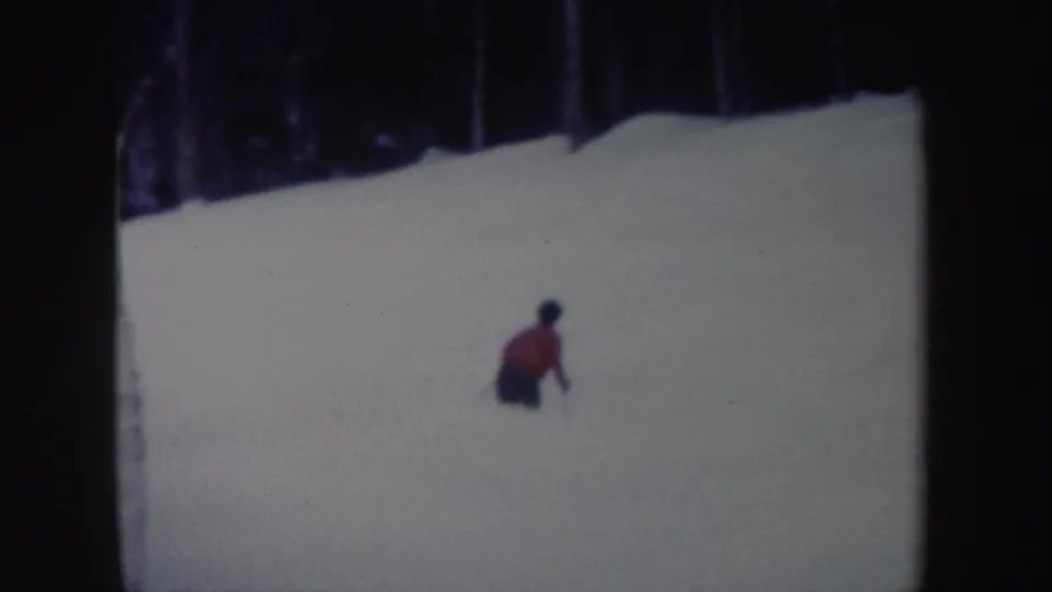 A red-jacketed skier crouches into a turn on the snowy New York slope in soft 8mm color footage.