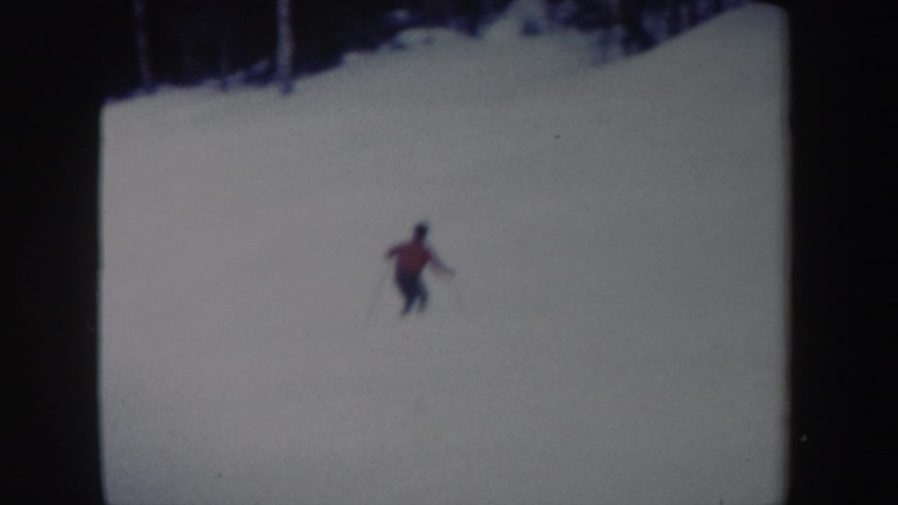 The skier is seen farther away on the open snow, descending below a dark treeline in 1962 home-movie footage.