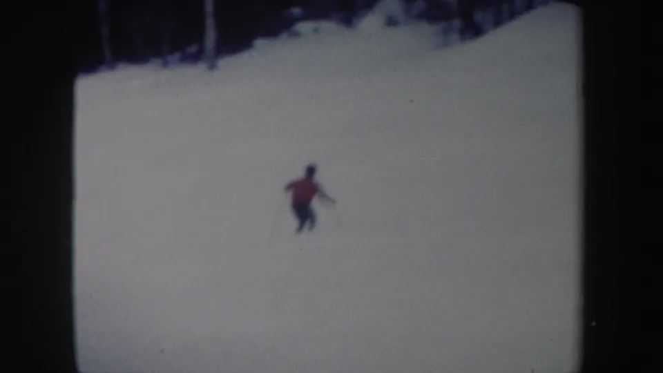 The skier is seen farther away on the open snow, descending below a dark treeline in 1962 home-movie footage.