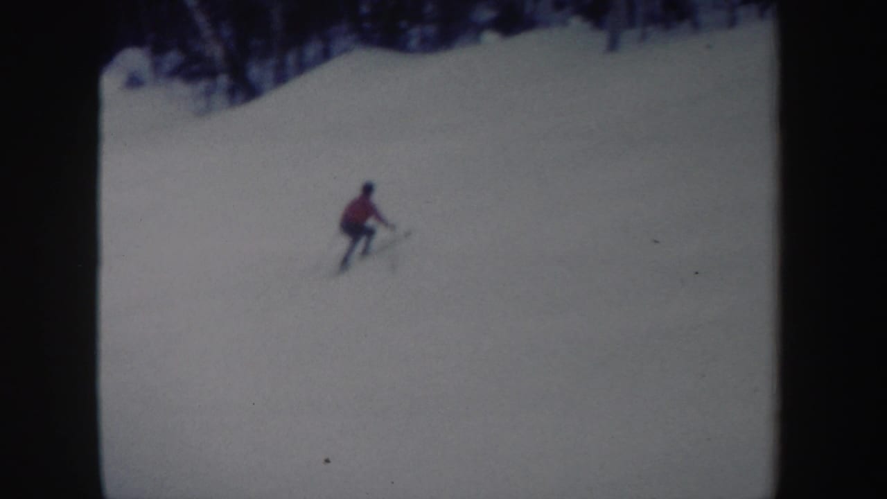 1962 New York 8mm frame showing a distant skier in red moving across a snowy hill with trees in the background.