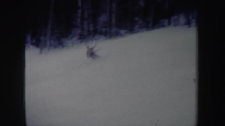 1962 New York home-movie frame with a small red-shirted skier descending a wide snow-covered slope near dark trees.