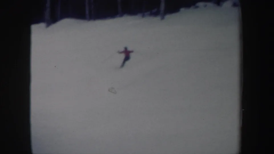1962 New York home-movie frame of a red-shirted skier gliding across a bright snowy hill below a dark wooded edge.