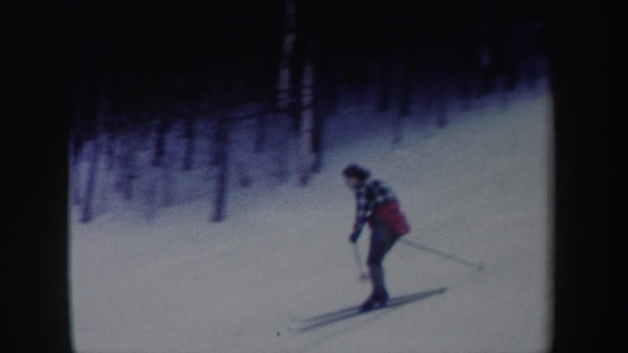 In 1962 New York home-movie footage, a woman in a plaid jacket and red garment skis on a snowy slope bordered by trees.