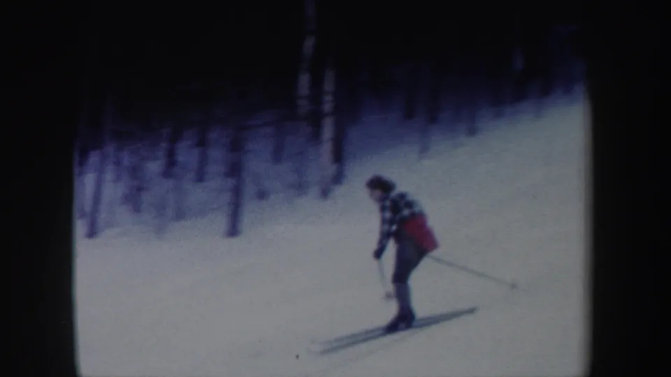 In 1962 New York home-movie footage, a woman in a plaid jacket and red garment skis on a snowy slope bordered by trees.
