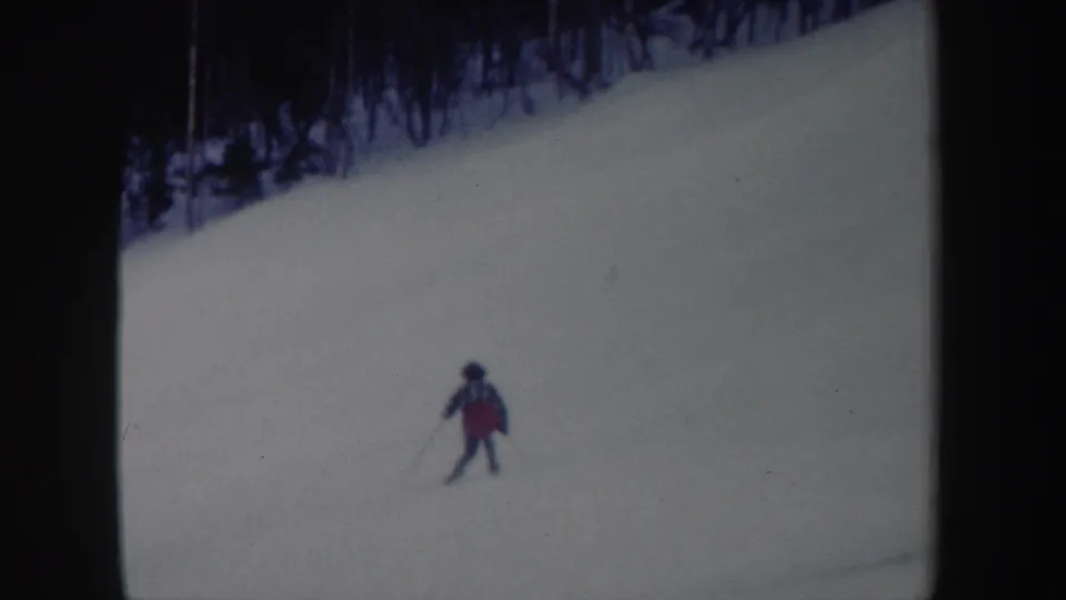 The skier appears smaller in frame as she continues down the snowy slope, with dark trees lining the upper edge.