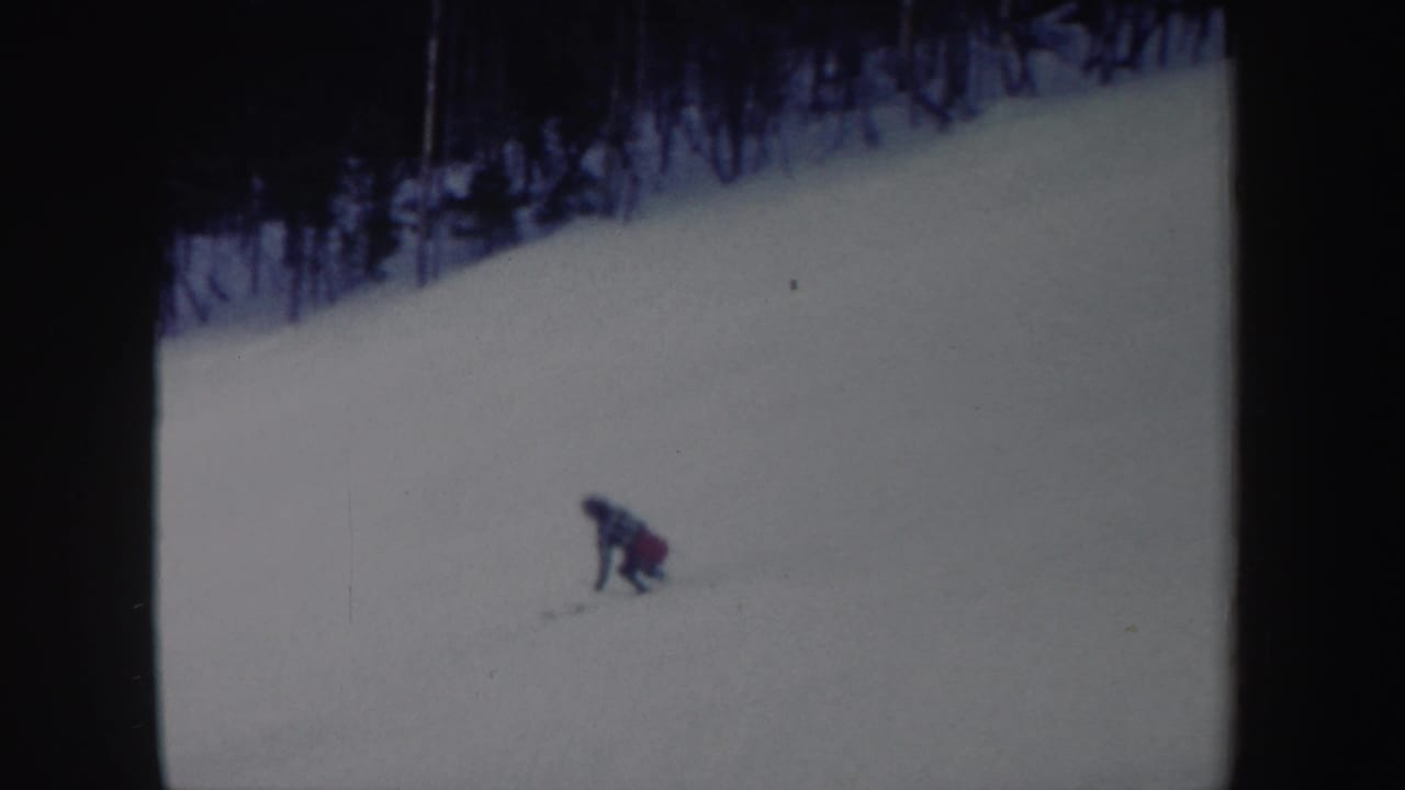 Lone skier in red and dark clothing bends on a snowy slope near a dark treeline.