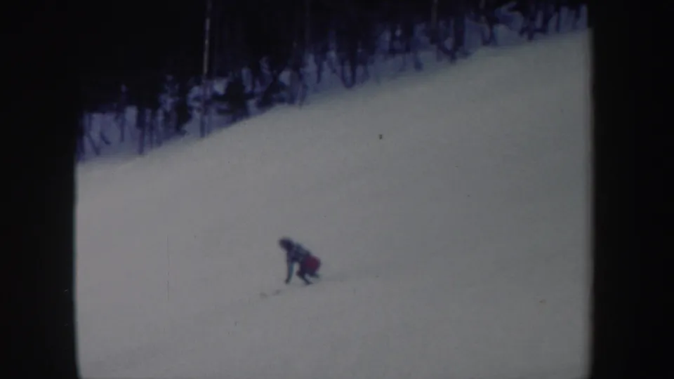 1962 New York 8mm frame showing a distant skier leaning low while turning on a snowy slope bordered by woods.