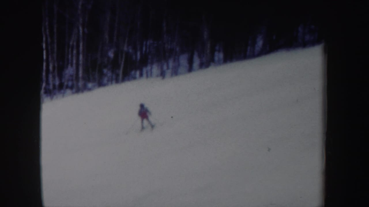 1962 New York home-movie frame showing a lone skier descending a snowy slope below a line of bare winter trees.