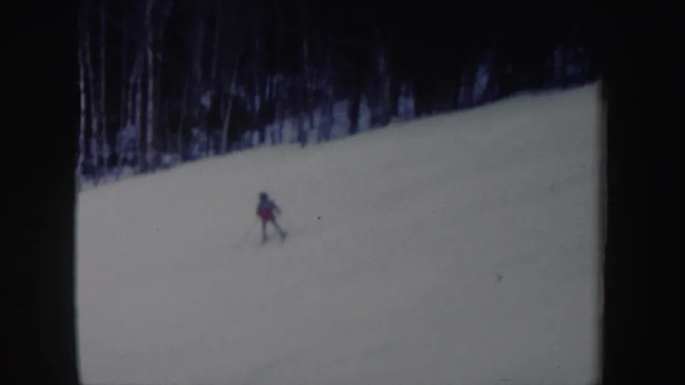 1962 New York home-movie frame showing a lone skier descending a snowy slope below a line of bare winter trees.