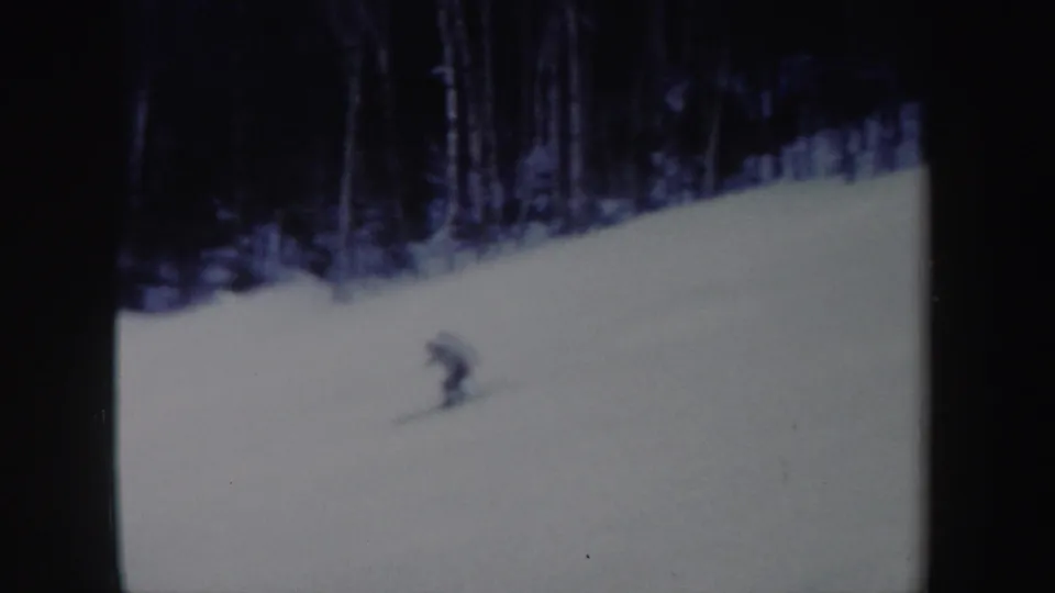 A distant skier continues downhill on a snowy New York slope, framed against a dark winter tree line in 1962 home-movie footage.