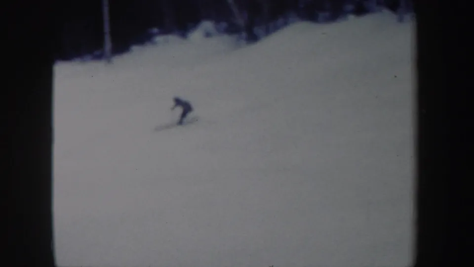 Distant 8mm frame of a lone skier leaning into a descent on a broad snowy slope in New York, 1962.