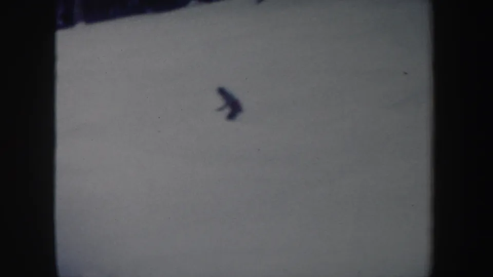 A lone skier is seen at long distance on a snowy slope, with soft 8mm grain and minimal background detail.