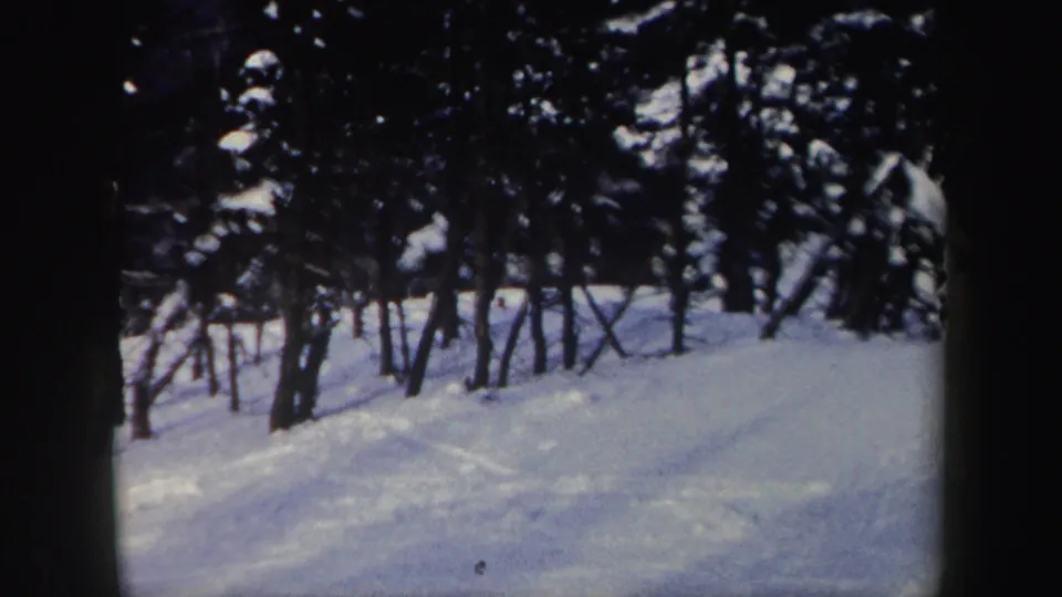 A wooded New York ski slope fills the frame, with snow tracks and dark trees visible in the 1962 home-movie footage.
