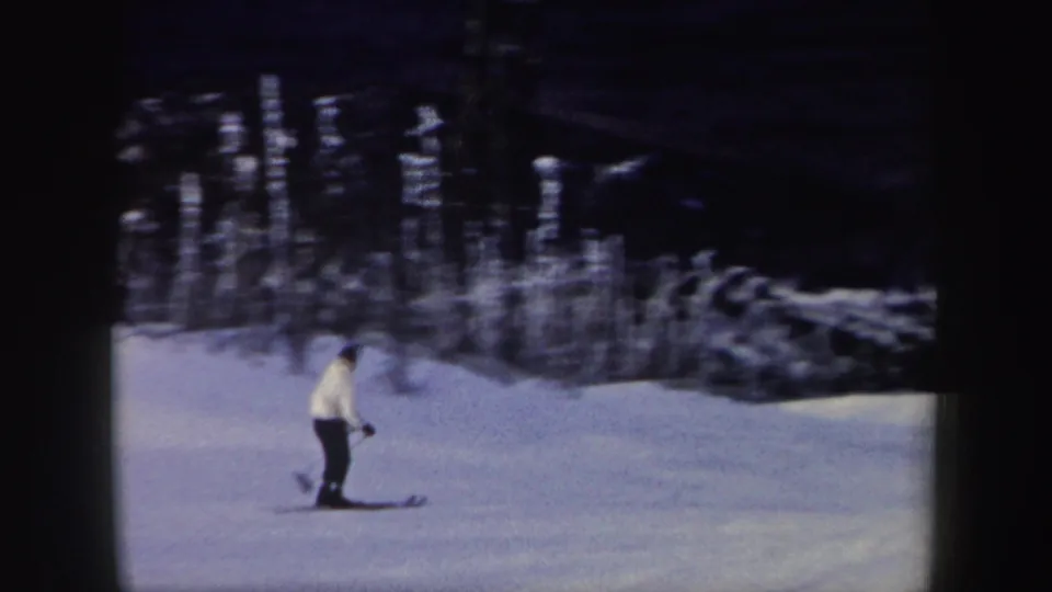 A distant skier in a light jacket and dark pants moves across a snowy New York hillside in 1962 home-movie footage.