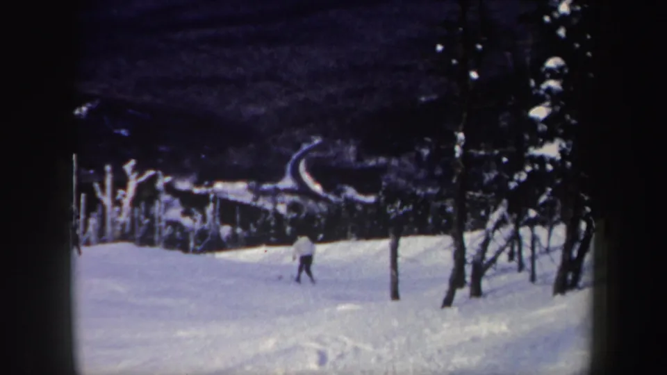 A small figure on skis descends farther down the snowy New York hill, with dark woods and visible tracks behind them.