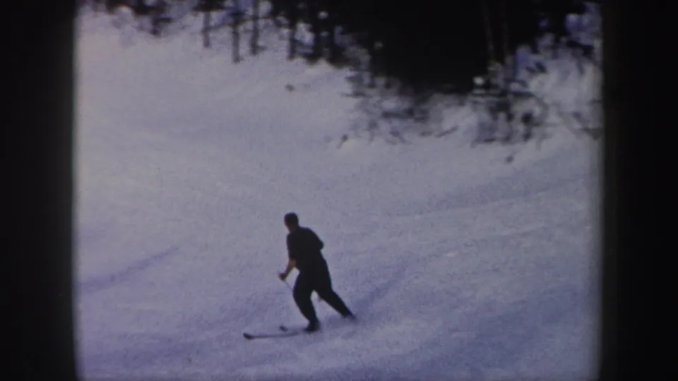 In this 1962 New York 8mm frame, a distant skier in dark clothing uses poles while crossing a snowy slope below a tree line.