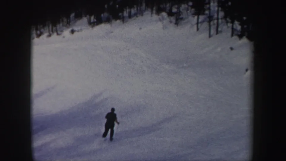 A lone skier appears small against a broad snowy New York slope, with trees along the upper edge of the 8mm frame.