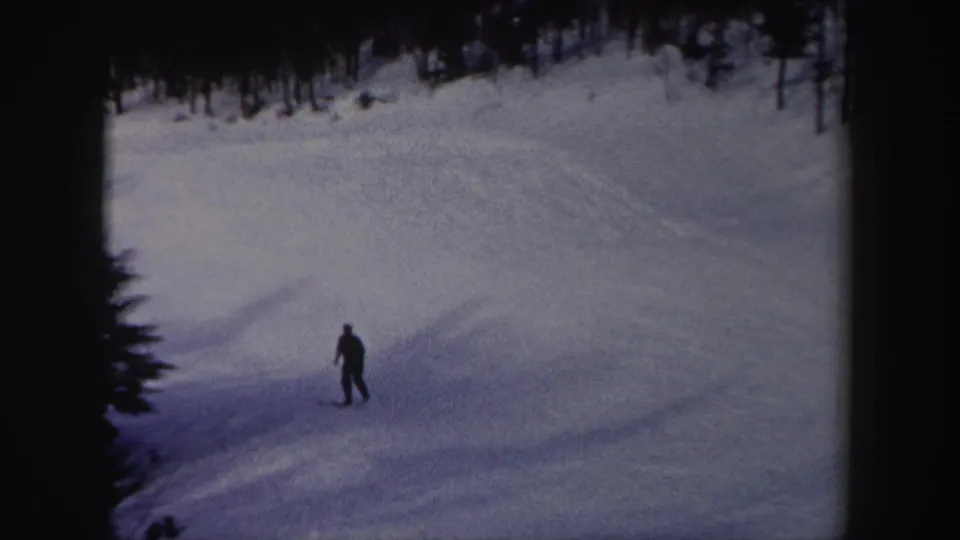 The skier is seen farther downhill on the snowy slope, framed by dark trees and soft 8mm film texture.