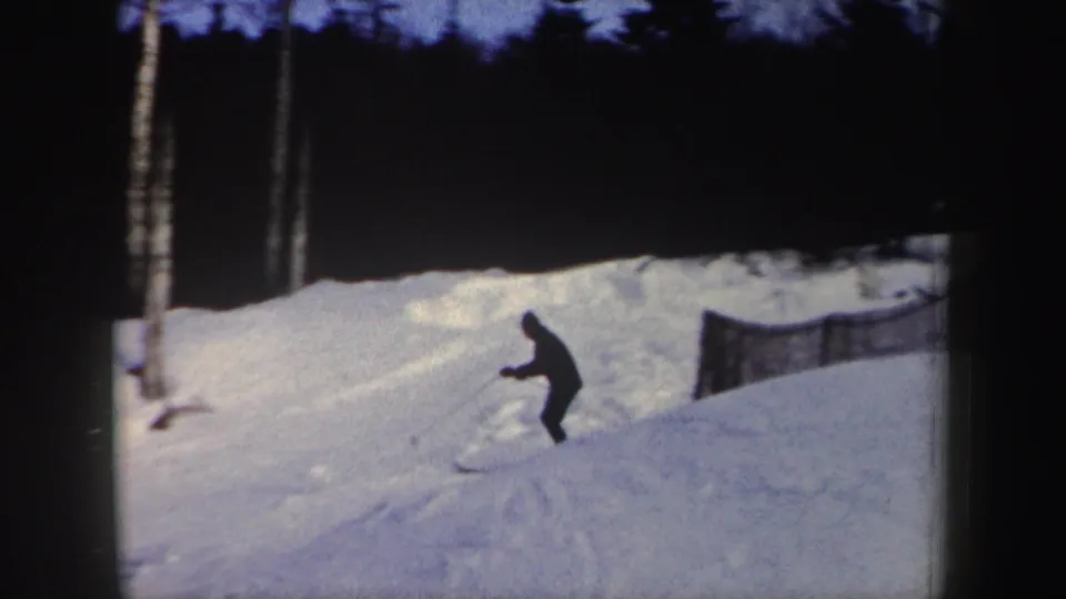 A skier crouches toward a small snow ramp on a wooded New York slope in metadata-supported 1962 8mm footage.