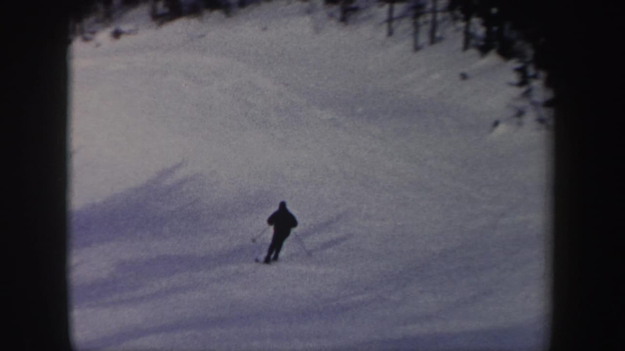 A distant skier descends a wide snowy run, with the handheld 8mm frame emphasizing open snow and forest edges.