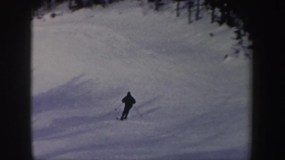 A distant skier descends a wide snowy run, with the handheld 8mm frame emphasizing open snow and forest edges.