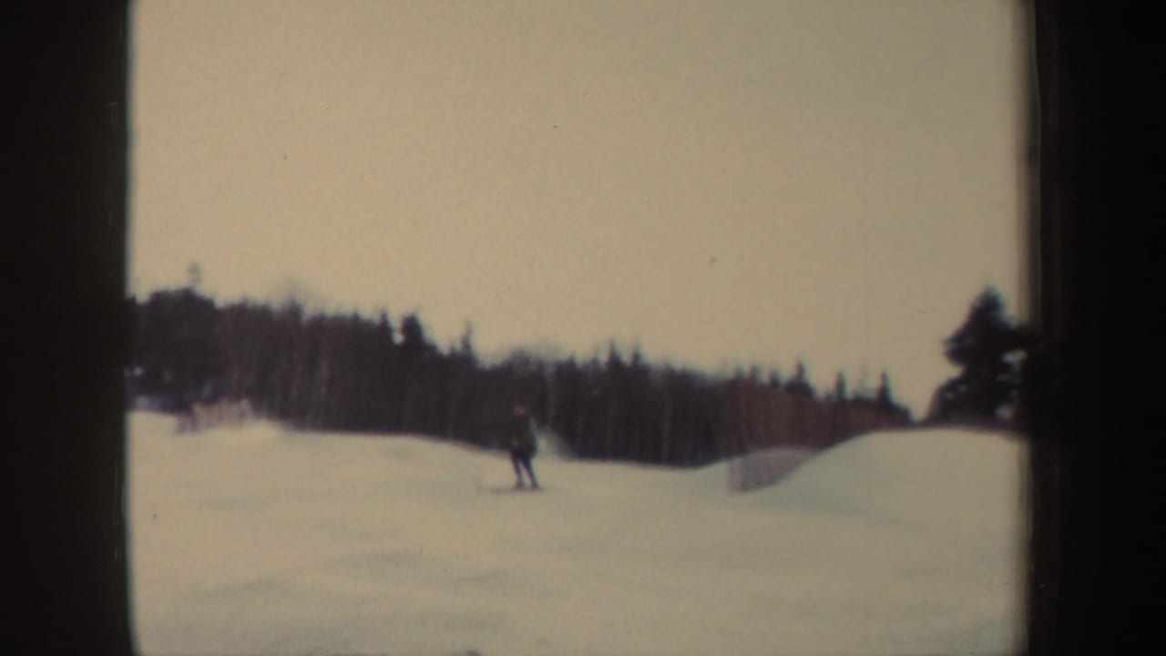 Distant skier crossing a snowy slope with a dark wooded tree line behind him.