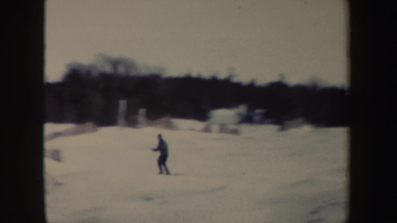 A lone skier in dark clothing moves across a snowy New York slope, with trees and snowbanks behind him.