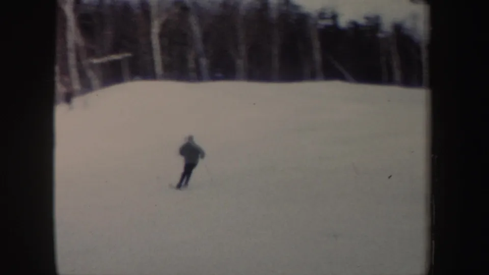 A distant skier crosses a broad snowy slope in New York, with winter trees along the upper edge of the frame.