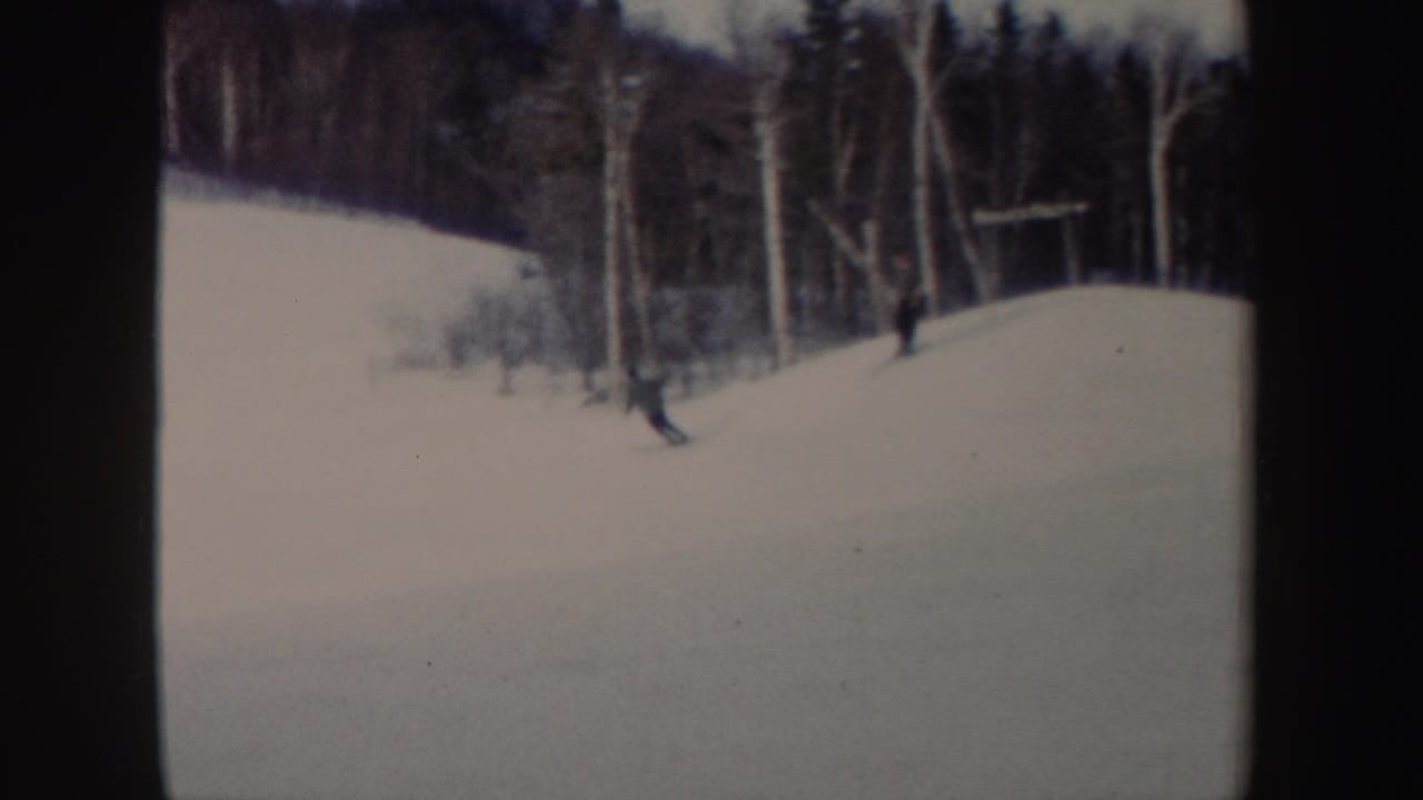 A skier passes near the edge of a snowy hill, with bare trees and a second distant figure or shadow-like form in the background.