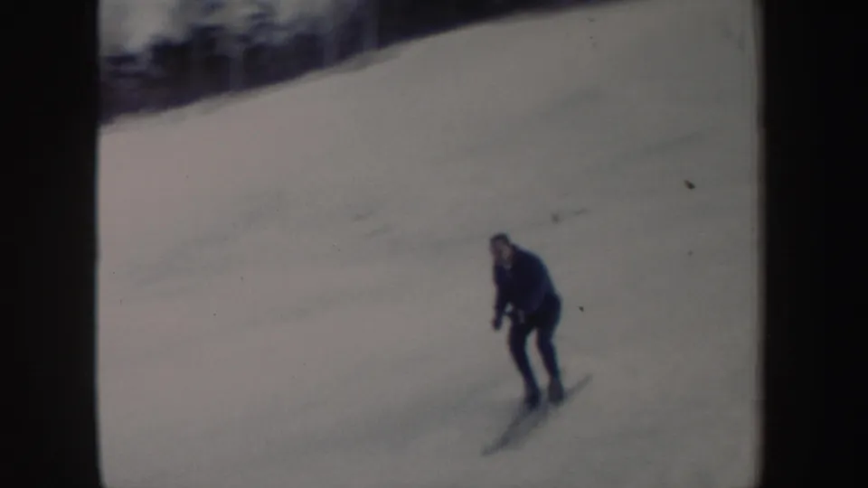 Adult skier in a dark blue coat makes a turn on a New York snowy slope in 1962 home-movie footage.