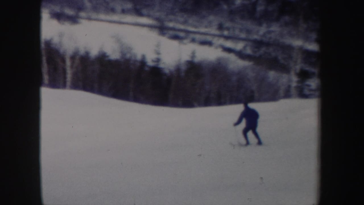 A distant skier crosses an open snowy slope with a tree line in the background in 1962 New York ski footage.