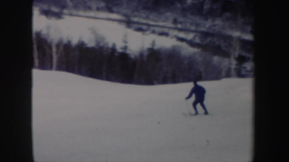 A distant skier crosses an open snowy slope with a tree line in the background in 1962 New York ski footage.