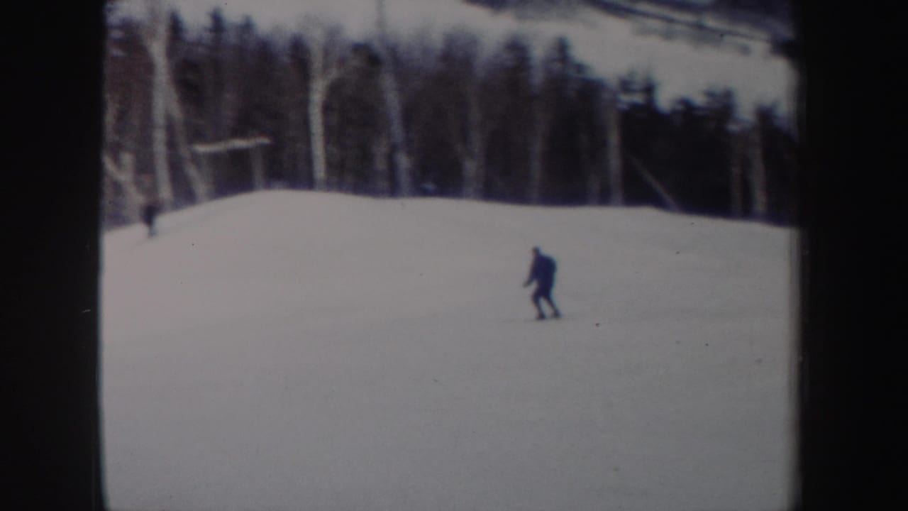 A lone skier continues downhill across a broad snowfield, with trees and possible slope equipment in the background.