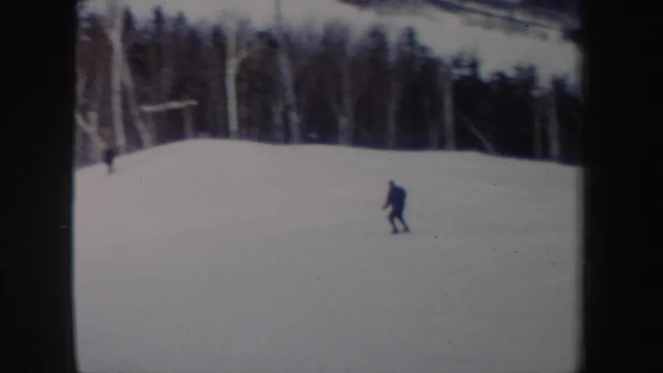 A lone skier continues downhill across a broad snowfield, with trees and possible slope equipment in the background.