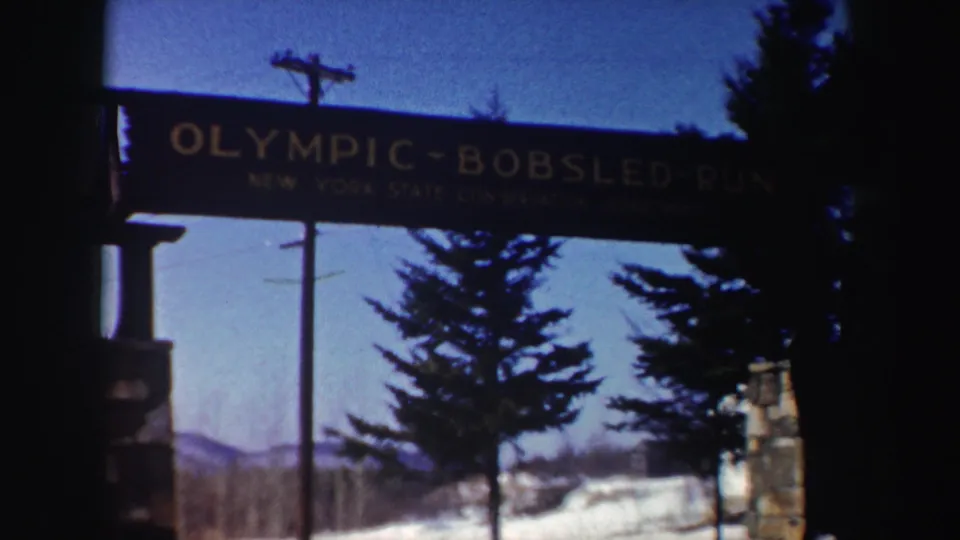 1962 New York 8mm frame of the Olympic-Bobsled-Run entrance sign above snowbanks, evergreens, stone posts, and a clear winter sky.