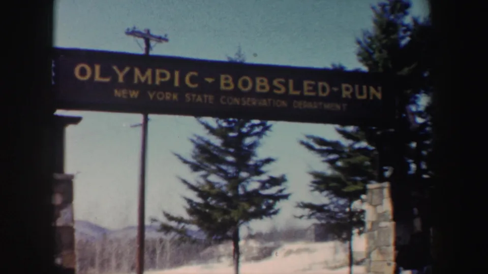 1962 New York 8mm frame of the Olympic Bobsled Run sign with snowbanks, conifers, stone posts, and mountains beyond.