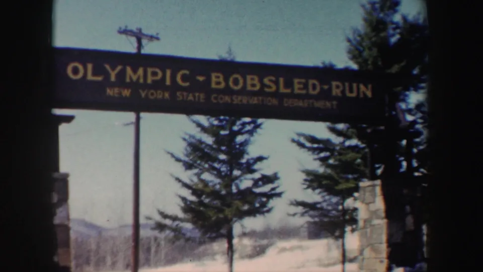 Final 1962 New York frame holds on the Olympic Bobsled Run entrance sign, stone supports, snow, evergreens, and mountain backdrop.