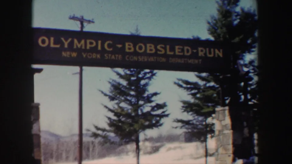 1962 New York home-movie frame with the Olympic Bobsled Run sign, snowy ground, trees, and a stone entrance post.