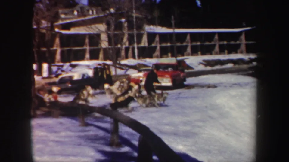 A man in a dark sweater or jacket rides behind a dog team on snowy ground in the 1962 New York home-movie footage.