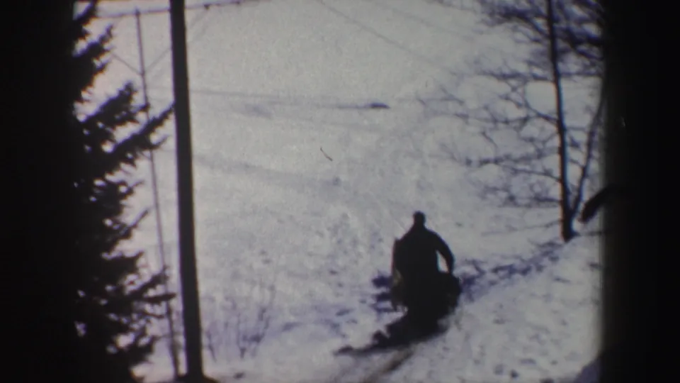 A distant musher rides behind a sled dog team on a snowy New York trail in a grainy 1962 8mm home-movie frame.