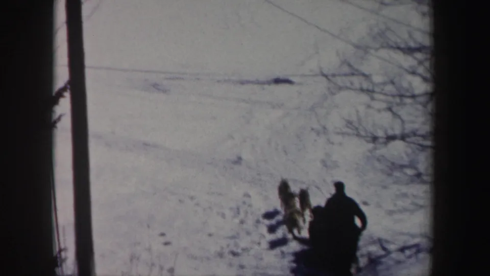 In 1962 New York home-movie footage, a silhouetted musher and dog team move across bright snow from a fixed viewpoint.
