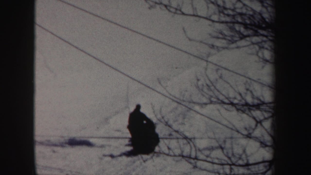 Distant bundled person pulls or guides a dark sled shape across snow.