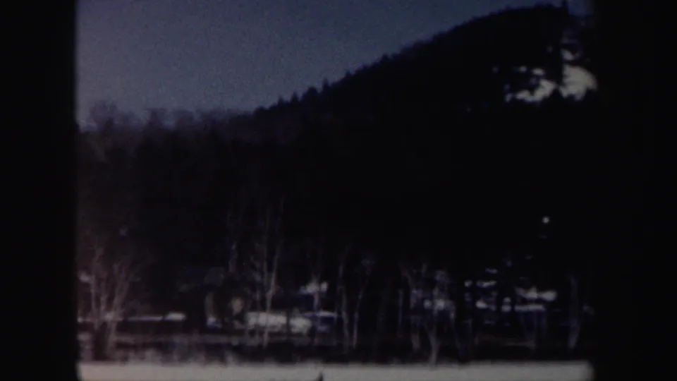 The 8mm frame shifts toward a dark wooded hillside with snowy ground, bare trees, and faint rural structures in the distance.