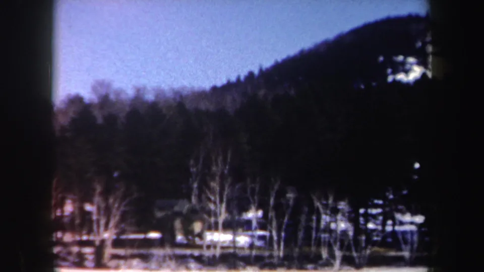 A snowy New York rural view shows bare trees, a dark wooded hill, and small distant buildings in soft, grainy 8mm color.
