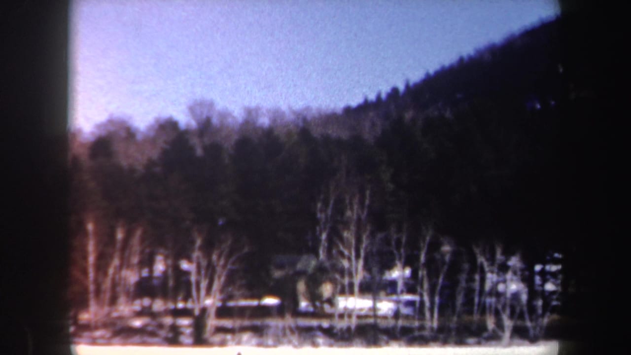 1962 New York 8mm frame showing a snow-covered lake edge, leafless trees, evergreens, and a wooded hillside.