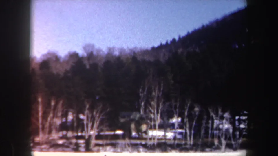 1962 New York 8mm frame showing a snow-covered lake edge, leafless trees, evergreens, and a wooded hillside.