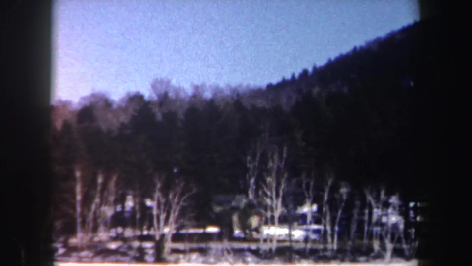 1962 New York home-movie frame looking across a snow-covered lake toward bare trees and a dark evergreen hillside.